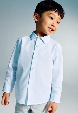 Young boy wearing a light blue button-up shirt and light pants, standing and looking to the side with a slight smile against a plain background.