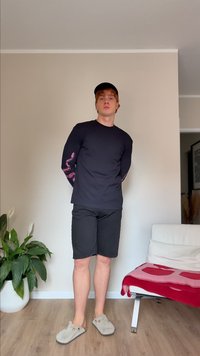 Camisa preta de manga longa com gráfico rosa nas mangas, combinada com calções pretos e sapatilhas cinzas. A pessoa está de pé em frente a um fundo neutro.