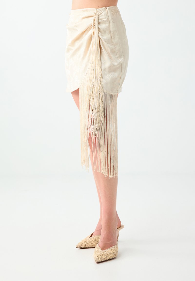 Mini-jupe beige avec un long détail à franges sur un côté, portée avec des escarpins beige tissés à bout pointu.