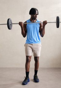 Sportler in einem hellblauen Ärmellosen Oberteil und beigen Shorts, der eine Langhantel mit schwarzen Gewichten hebt. Strukturierte schwarze Sportschuhe und dunkle Socken.