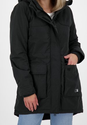Femme portant un manteau d'hiver noir à capuche avec des poches avant, dézippé sur une chemise rayée et un jean bleu clair.