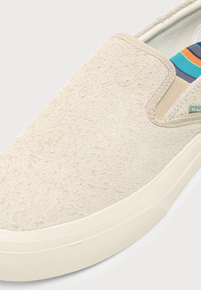 Μπεζ παπούτσι τύπου slip-on με υφασμάτινο επάνω μέρος με υφή, ελαστική πλαϊνή πλευρά και λευκή λαστιχένια σόλα, που δείχνει πολύχρωμη εσωτερική επένδυση.
