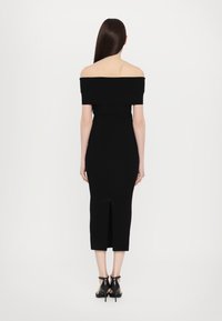 Black off-the-shoulder dress with a fitted silhouette, knee-length hem, back slit opening, and smooth texture. Heeled black shoes visible.