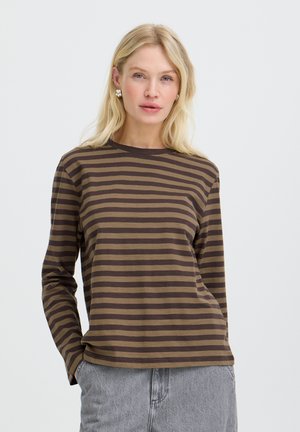 Lyshåret kvinde iført brun og sort stribet langærmet skjorte og lysegrå jeans, stående med den ene hånd i lommen.