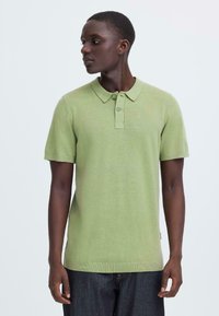 Polo à manches courtes en tissu maille vert clair avec patte de boutonnage à trois boutons, col et ourlet côtelé. Porté avec un jean denim foncé.