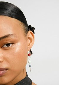 Close-up van een vrouw met stijlvol zwart haar, die een lange oorring draagt met rode, witte en blauwe kralen en zilveren bedels.