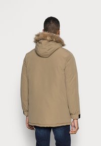 Muž otočený zády, oblečený v béžové zimní bundě s koženým lemem a tmavě modrých džínách, na jednolitém světlém pozadí.