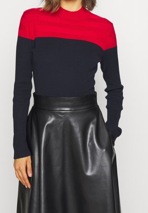 Femme portant un pull à manches longues côtelé rouge et bleu marine, rentré dans une jupe A-line en cuir noir taille haute avec poches.