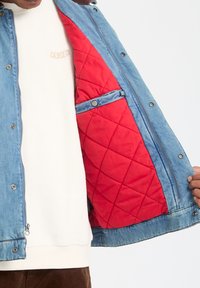 Denim jacket met een rode gewatteerde voering, voorzien van drukknoop- en ritssluitingen. De buitenkant is lichtblauw en heeft een gladde textuur.