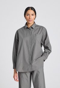 Camicia grigia button-up con maniche lunghe, in tessuto di cotone, con taschino sul petto, vestibilità comoda e una texture sottile. Indossata con pantaloni grigi abbinati.