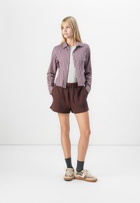 Kvinde stående med hænderne i lommerne iført en ternet skjorte, grå top, brune shorts med logo, mørke sokker og mønstrede sneakers på hvid baggrund.