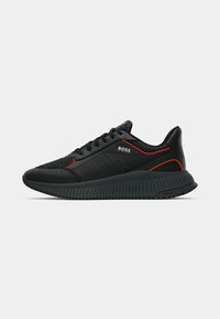 Chaussure de sport noire avec une tige en mesh et cuir, dotée d'une semelle texturée, d'accents rouges sur les côtés, et d'un logo sur la languette et le talon.