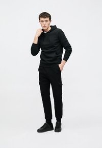 Felpa con cappuccio in cotone nero con coulisse, abbinata a pantaloni cargo jogger neri con tasche laterali. Indossata con sneakers nere, design minimalista.