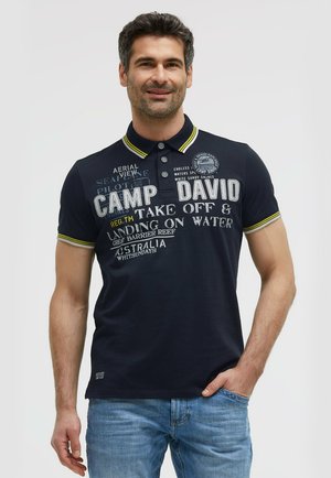 Man with dark hair wearing a navy polo shirt with yellow trim and white text, standing with one hand in his blue jeans pocket.