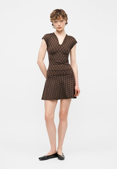 Νεαρή γυναίκα που φοράει καφέ φόρεμα με μοτίβο, κοντά μανίκια και V-λαιμόκοψη, καθώς και μαύρες μπαλαρίνες, στέκεται μπροστά σε λευκό απλό φόντο.