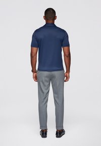 Granatowa koszulka polo z krótkim rękawem, zestawiona z jasno szarymi, taliowanymi spodniami. Model stoi tyłem, prezentując zaokrąglony dół koszulki i podwinięte nogawki spodni.
