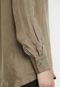 Olivově zelená košile s dlouhým rukávem z lehkého materiálu, s nařasenými detaily na rukávech a knoflíkem na manžetě.