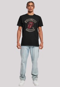 Zwarte katoenen T-shirt met een opvallend rood tonglogo, "Rolling Stones"-tekst en een losse pasvorm. Gekocht met lichtblauwe jeans en witte sneakers.