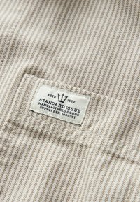 Gros plan sur un tissu beige à rayures avec une étiquette blanche cousue indiquant « Standard Issue Manufactured Goods ESTD 1982 Supply Ref: 100578T. »