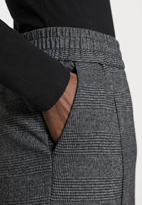 Grå rutete bukser med elastisk liv og sidelommer. Stoffet har et strukturert mønster som øker visuell interesse.