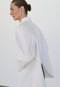 Camisa blanca de gran tamaño con hombros estructurados, que presenta un diseño de espalda partida. Tejido suave con detalles mínimos y una silueta limpia.