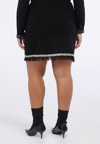 Schwarzes Kleid mit schlichtem Design, verziert mit einem silbernen Fransen-Saum, kombiniert mit schwarzen Kniestrümpfen und glänzenden schwarzen High Heels.