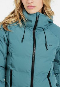 Türkiser Pufferjacke mit hohem Kragen, schwarzem Reißverschluss, verstellbaren Kordeln und seitlichen Taschen. Glatte Textur und moderne Designdetails.