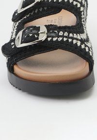 Sandale tissée noir et blanc avec deux boucles argentées ajustables, semelle en tan et semelle extérieure en caoutchouc noir texturé.