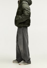 Chaqueta acolchada verde oliva con capucha, combinada con jeans gris claro anchos y zapatillas grises con cordones blancos. Look casual de streetwear.