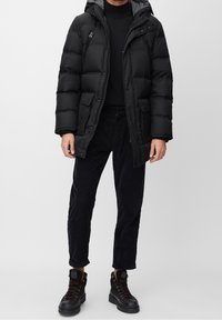 Chaqueta de plumas negra con capucha, que presenta un cierre de cremallera frontal, dos bolsillos frontales y un diseño acolchado. Lleva puesta con pantalones de pana negros y botas.