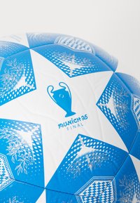 White soccer ball with blue accents, featuring a geometric pattern, trophy logo, and "Munich 25 Final" text. Smooth texture, stitched panels.