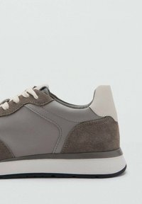 Sneaker grigia con una combinazione di materiali sintetici lisci e scamosciati. Presenta lacci grigio chiaro, un tallone testurizzato e una suola scura.