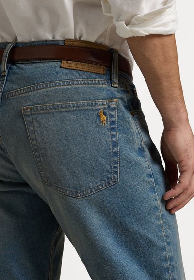 Heled sinised teksad on sirge lõikega, pruunist nahast vöökoht ja eristuv kollane polo logo tagakotis.