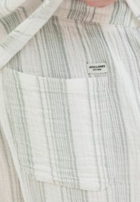 Camisa a rayas blanca y verde claro con un bolsillo horizontal y una pequeña etiqueta de marca, hecha de un tejido texturizado y ligero.