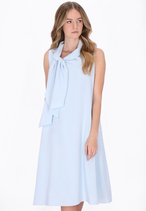 Młoda kobieta z długimi falującymi włosami, ubrana w bezrękawową, jasnoniebieską sukienkę do kolan z dużą, związaną kokardą przy dekolcie.