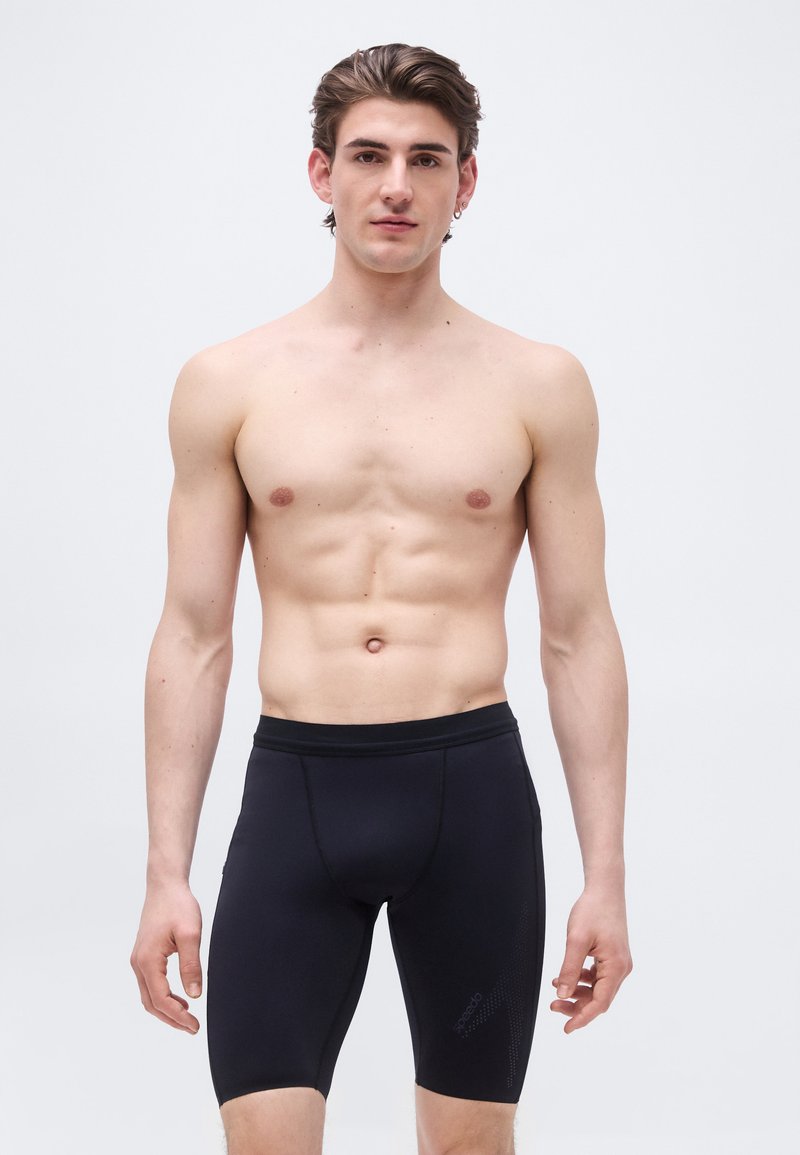 Γυμνόστηθος νεαρός με κοντά καστανά μαλλιά, που φοράει στενά μαύρα μαγιό μέχρι το γόνατο, στέκεται μπροστά σε απλό φωτεινό φόντο.