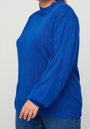 Femme portant un pull bleu roi à manches longues avec de légères fronces aux épaules et un jean bleu, montrée de la moitié du torse au cou et à la main.