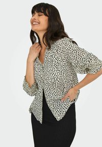 Button-up blouse in lichte, crèmekleurige stof met zwart gespikkeld patroon; heeft pofmouwen en een ronde zoom. Gecombineerd met een zwarte rok.