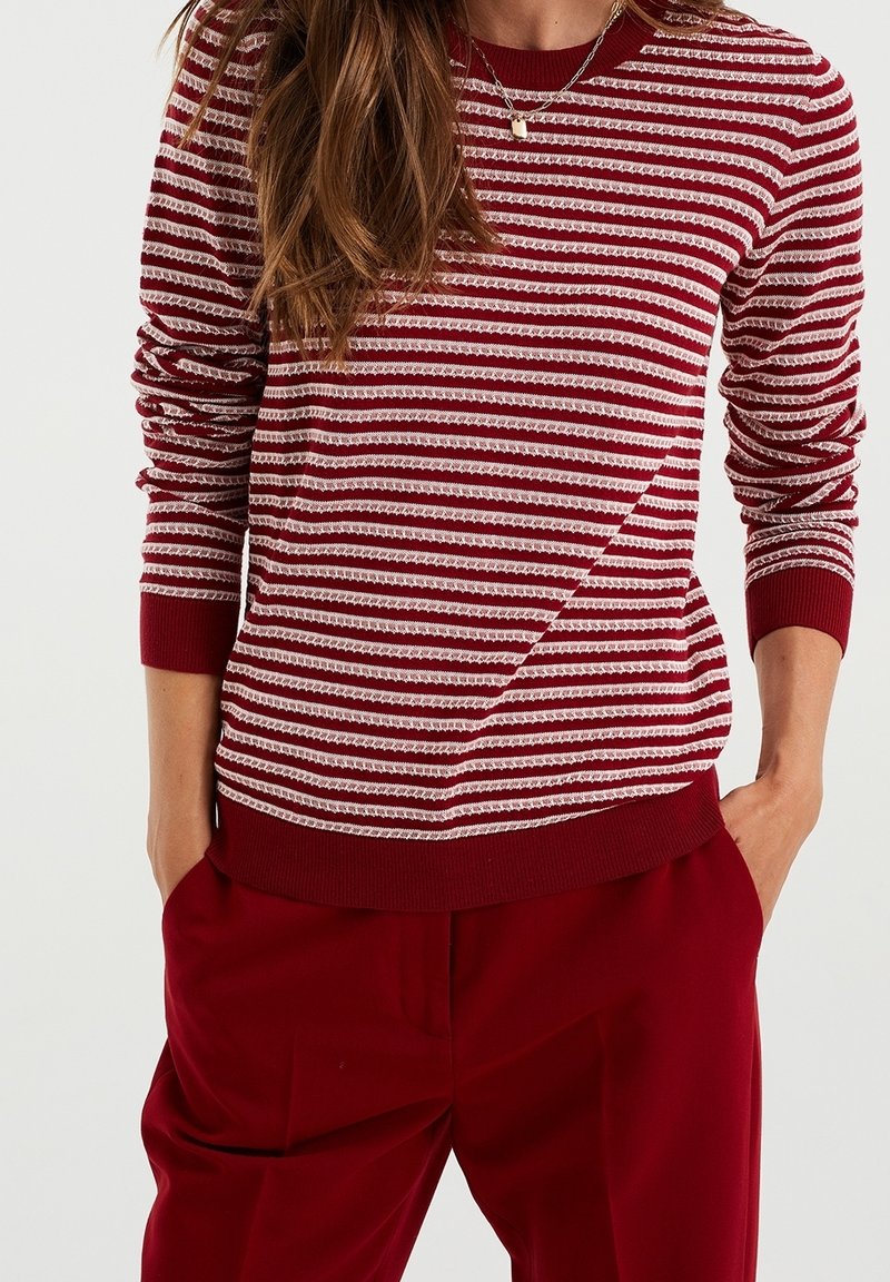 Femme portant un pull à manches longues rayé rouge et blanc avec un tricot texturé, assorti à un pantalon rouge uni, mains dans les poches, collier visible.