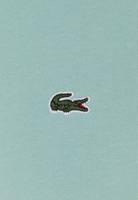 Tessuto di cotone verde menta chiaro con un piccolo logo di coccodrillo ricamato in verde, con contorno bianco e dettagli rossi sul muso.