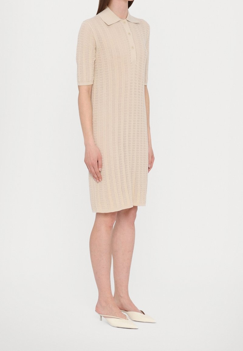 Mannequin féminin portant une robe polo en maille beige avec des manches jusqu'au coude et des mules crème à talons bas pointues, debout sur un fond uni.