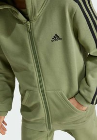 Sudadera verde con cremallera y acabado texturizado, con franjas negras en los lados y un logotipo de Adidas en el pecho. Incluye dos bolsillos delanteros.
