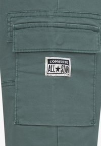 Zöld cargo nadrág, amelyen egy nagy, hajtókás zseb található, a Converse All Star címkével, amely egy kontrasztos fehér folton van hímezve. Puha szövet textúra.
