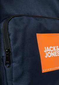 Mochila de tela azul marino con un prominente logo rectangular naranja con texto blanco. Incluye un zipper negro en un lado. Material texturizado.