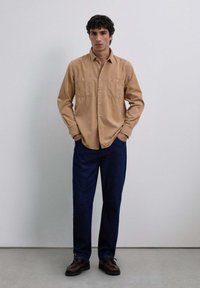 Beżowa koszula zapinana na guziki z dwoma kieszeniami na piersiach, w zestawieniu z ciemnoniebieskimi spodniami i brązowymi butami. Model stoi z rękami w kieszeniach na neutralnym tle.