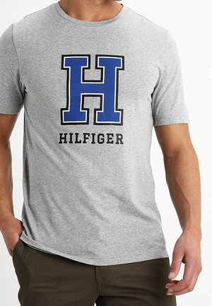 Šedé tričko s krátkým rukávem, na kterém je velké modré písmeno "H" a černé slovo "HILFIGER", vyrobené z měkkého bavlněného materiálu.