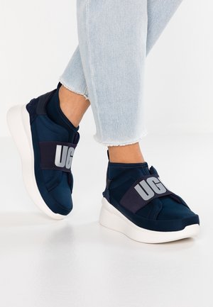 Füße in dunkelblauen Slip-on-Sneakern mit breiten elastischen Riemen mit der Aufschrift "UGG" und hellblauen Jeans mit ausgefranstem Saum auf weißem Boden.