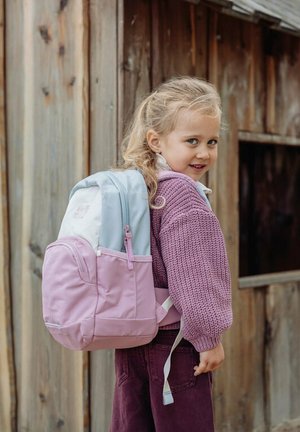 Jeune fille aux cheveux blonds portant un pull violet et un sac à dos rose, debout à l'extérieur près d'un bâtiment en bois, regardant en arrière et souriant.