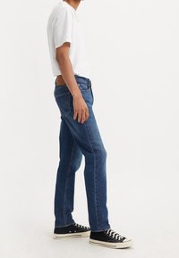 Blå denimjeans med raka ben, med en brun läderlapp på baksidan, bärs med en vanlig vit t-shirt och svarta sneakers.