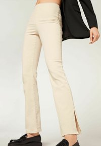 Pantaloni beige a gamba svasata con spacchi laterali, realizzati in tessuto morbido. Abbinati a mocassini neri, su uno sfondo chiaro e semplice.